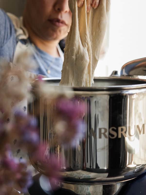 A person kneads dough with both hands in a shiny metal mixer. Flowers are blurred in the foreground, adding a touch of color to the scene. The focus is on the dough stretching out from the mixer.